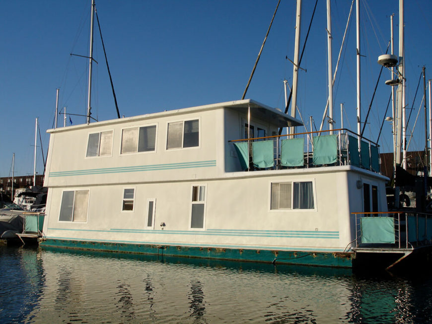 A large moored houseboat designed to be a permanent home. The boat has no side railings, and two small decks on the front and rear of the boat. Like many permanent house boats in North America, this one is moored in a large marina. A large moored houseboat designed to be a permanent home. The boat has no side railings, and two small decks on the front and rear of the boat. Like many permanent house boats in North America, this one is moored in a large marina.