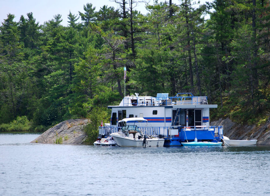 A massive houseboat with a variety of other boats moored to it off the coast of a forested island. This is a perfect example of house boating for leisurely purposes. A massive houseboat with a variety of other boats moored to it off the coast of a forested island. This is a perfect example of house boating for leisurely purposes.