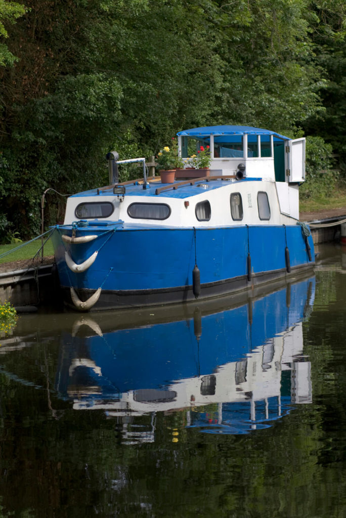A large houseboat in bright blue and contrasting white moored off of a canal. Unlike many other houseboats, this one has very few windows and no open A large houseboat in bright blue and contrasting white moored off of a canal. Unlike many other houseboats, this one has very few windows and no open
