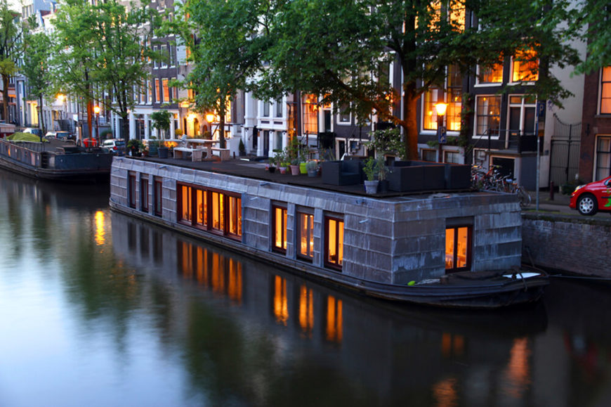 A large shingled houseboat in Amsterdam with a variety of potted plants and patio furniture on the roof. A large shingled houseboat in Amsterdam with a variety of potted plants and patio furniture on the roof.