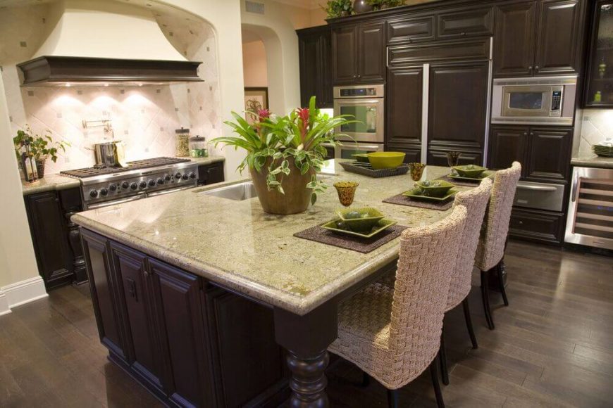 Dark brown cabinets are offset by a slightly paler brown floor while the off-white tile backsplash is complemented by the rough woven chairs standing attention at the island. Bright green accents are used around the room to bring in a pop of color to the otherwise dark space. Dark brown cabinets are offset by a slightly paler brown floor while the off-white tile backsplash is complemented by the rough woven chairs standing attention at the island. Bright green accents are used around the room to bring in a pop of color to the otherwise dark space.