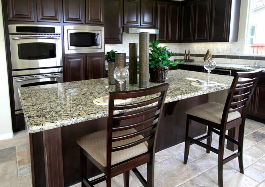 Pale counters, floors, and tile backsplash balance out the dark wood used throughout this kitchen. Bold textures offset the sleek, structured look of the cabinets and chairs. The introduction of greenery on the counter brings life and a splash of color into the space. Pale counters, floors, and tile backsplash balance out the dark wood used throughout this kitchen. Bold textures offset the sleek, structured look of the cabinets and chairs. The introduction of greenery on the counter brings life and a splash of color into the space.
