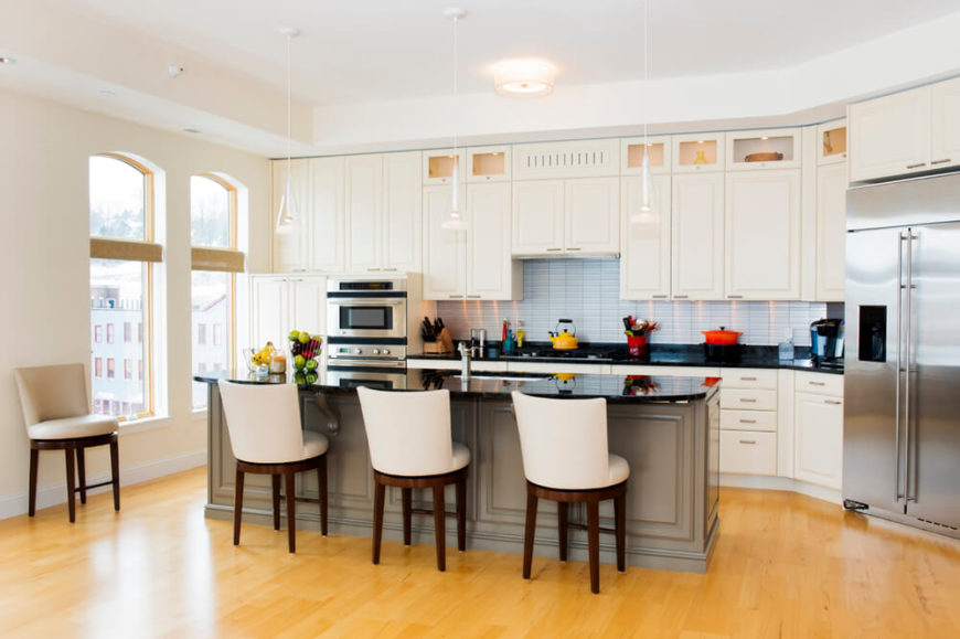 Creamy cabinets match the wall color in this open kitchen design. The designers used the bright white tiles of the backsplash to add a difference in color and a black countertop to break up the space. The warm gray of the island helps to differentiate it from the uniform counters. Creamy cabinets match the wall color in this open kitchen design. The designers used the bright white tiles of the backsplash to add a difference in color and a black countertop to break up the space. The warm gray of the island helps to differentiate it from the uniform counters.