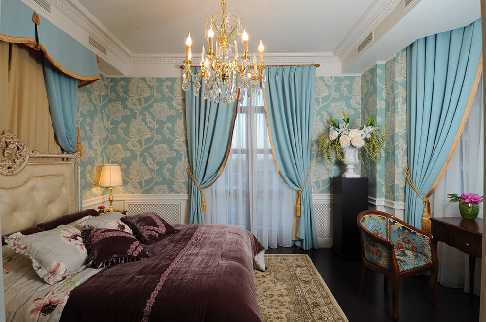 Charming primary bedroom clad in lovely blue floral wallpaper and lighted by a fancy gold chandelier that hung over the cream tufted bed dressed in mauve bedding.