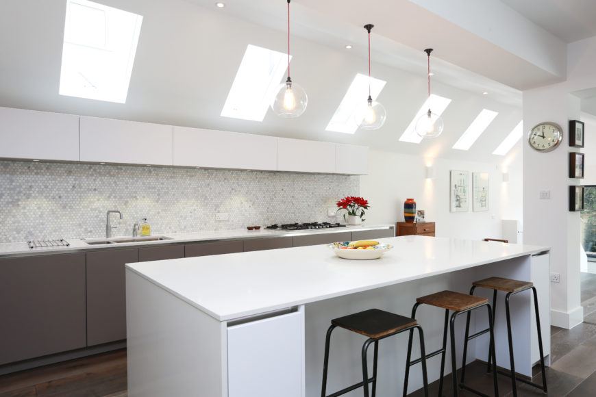 Modern kitchen island. Modern kitchen with white walls and center island along with tiles backsplash and a breakfast bar space lighted by pendant lights.