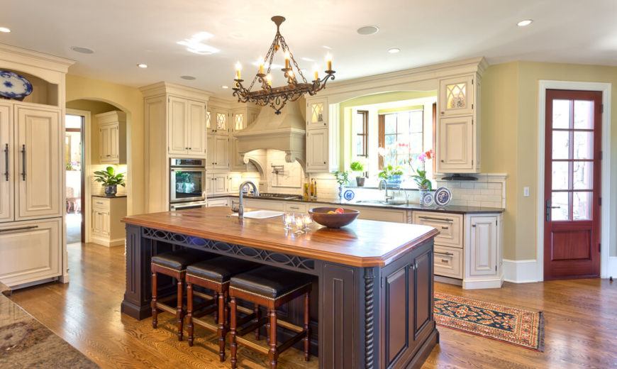 Huge kitchen with white cabinetry and hardwood floors along with a large center island featuring wooden countertop and space for a breakfast bar lighted by recessed lights and chandelier.