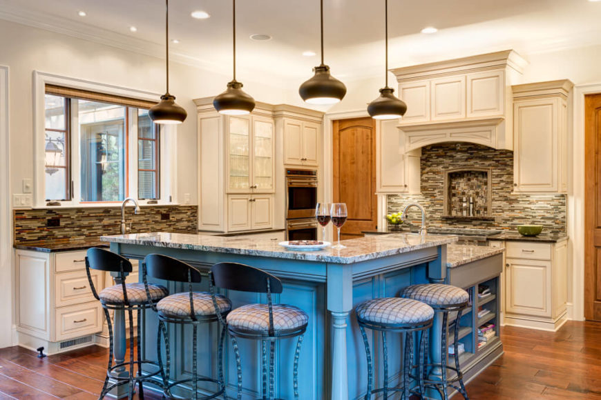 L shape kitchen with stylish camou backsplash and white cabinetry along with a center island featuring marble countertop and a breakfast bar lighted by recessed and pendant lights.