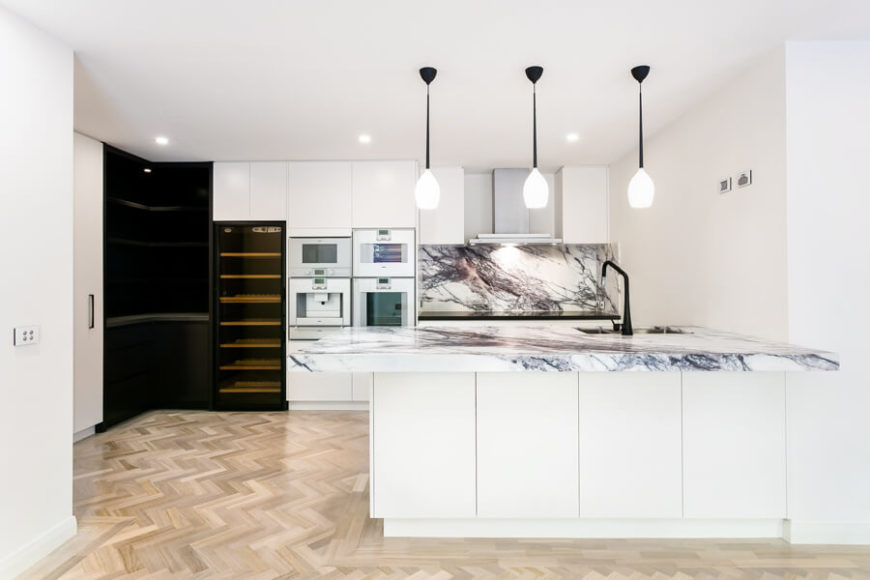Bright kitchen with white walls and marble countertops and backsplash lighted by recessed ceiling lights.