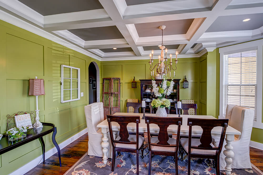 An olive green dining room showcases recessed wainscoting walls and coffered ceiling. It has a white dining table surrounded by cushioned wooden and skirted wingback chairs.