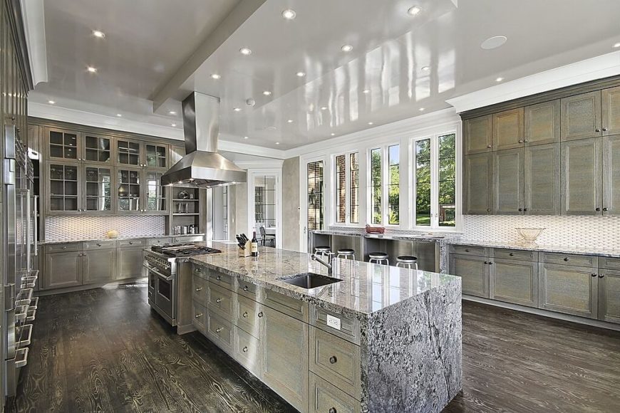 The massive, lengthy island covered in marble is the centerpiece of this vast kitchen, standing atop dark hardwood flooring and surrounded by rustic tinted cabinetry. The stainless steel appliances add a layer of sheen to the look without distracting from the overall style. The massive, lengthy island covered in marble is the centerpiece of this vast kitchen, standing atop dark hardwood flooring and surrounded by rustic tinted cabinetry. The stainless steel appliances add a layer of sheen to the look without distracting from the overall style.