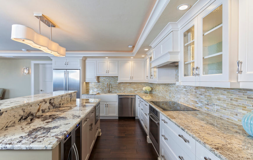 With a marble topped, two-tier island defining the edge of this kitchen, the stainless steel appliances help it stand out amid a large open plan home. Subtle tile backsplash complements the vast array of white cabinetry with steel hardware. With a marble topped, two-tier island defining the edge of this kitchen, the stainless steel appliances help it stand out amid a large open plan home. Subtle tile backsplash complements the vast array of white cabinetry with steel hardware.