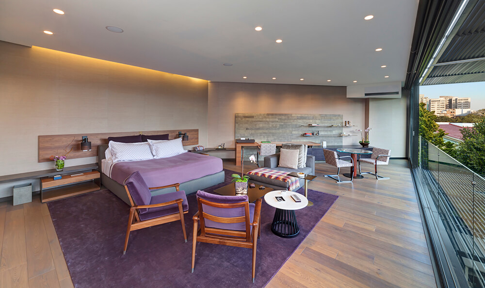 Huge primary bedroom with its own home office and a coffee table set. It features hardwood floors with a violet rug on top and is lighted by recessed ceiling lights.
