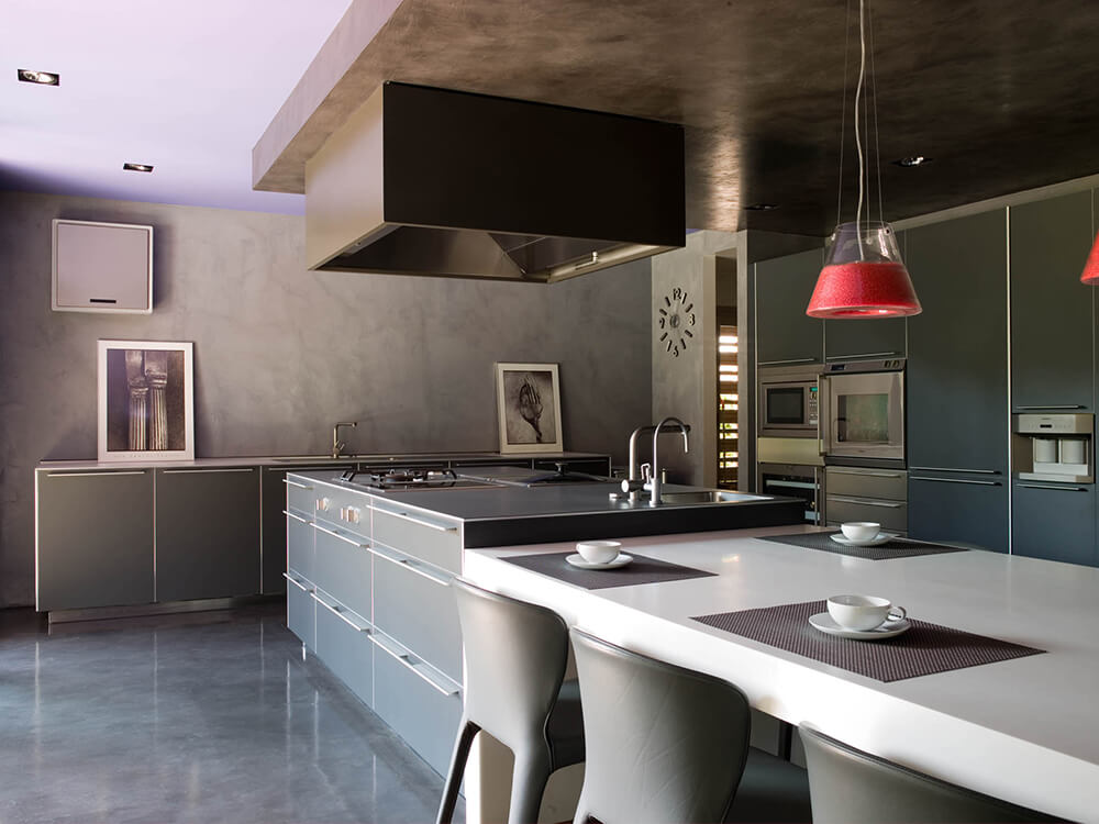 A modish industrial kitchen set with dark flooring and black kitchen counters and cabinets. The center island boasts a white breakfast bar on the side.