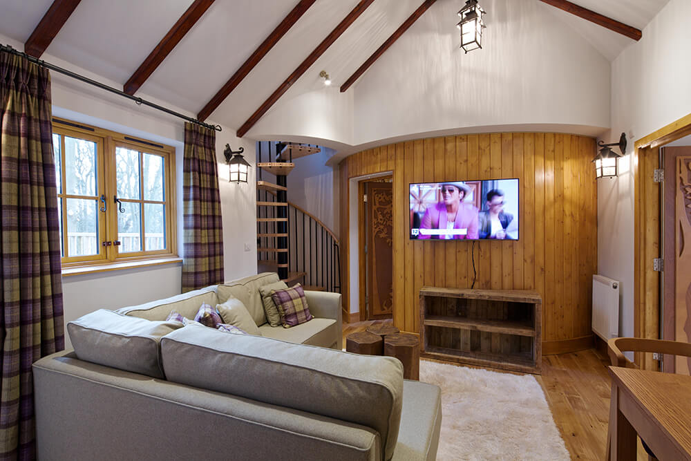 Craftsman living room offers wood plank flooring that complements with the accent wall mounted with a flat panel TV along with cathedral ceiling lined with dark wood beams.