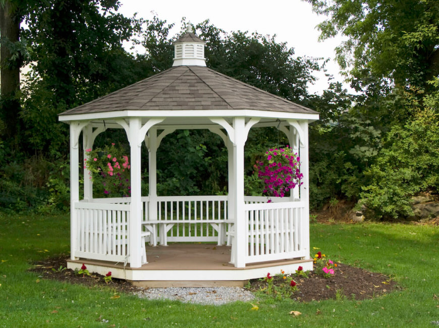 This white hexagonal gazebo features a small planting bed around it and brightly colored hanging baskets of flowers are displayed near the front. Built in benches provide seating. This white hexagonal gazebo features a small planting bed around it and brightly colored hanging baskets of flowers are displayed near the front. Built in benches provide seating.