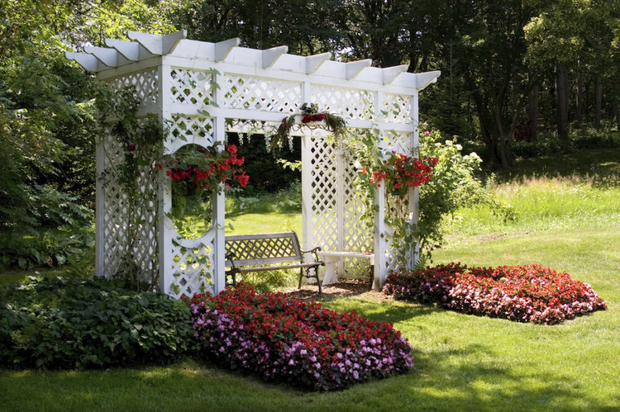 This white pergola is nestled above a small garden patio and bedecked with hanging baskets, climbing vines, and thick beds of flowers. The structure of the pergola is made up of trellises. This white pergola is nestled above a small garden patio and bedecked with hanging baskets, climbing vines, and thick beds of flowers. The structure of the pergola is made up of trellises.