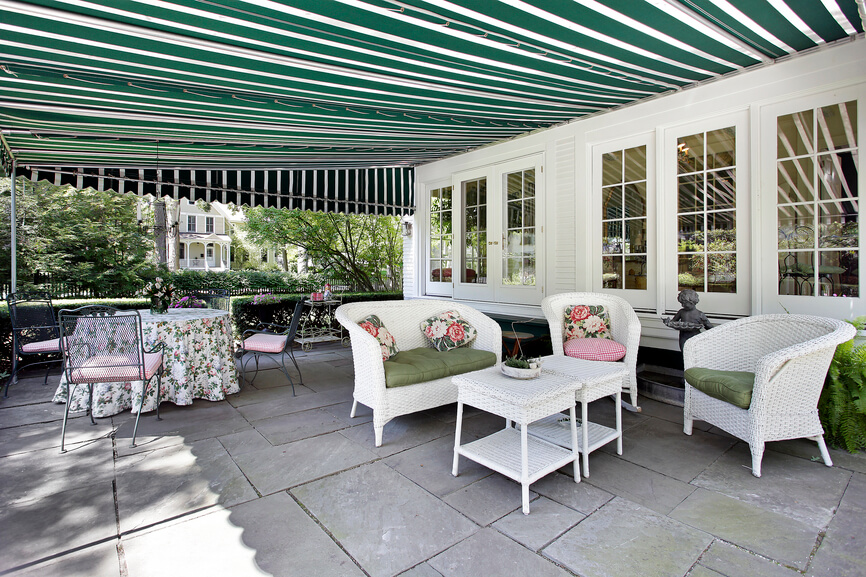 A shaded, quaint cottage style patio with large format flagstone tiles and a green striped awning. A hedge along one side hems in the relaxation area. A shaded, quaint cottage style patio with large format flagstone tiles and a green striped awning. A hedge along one side hems in the relaxation area.