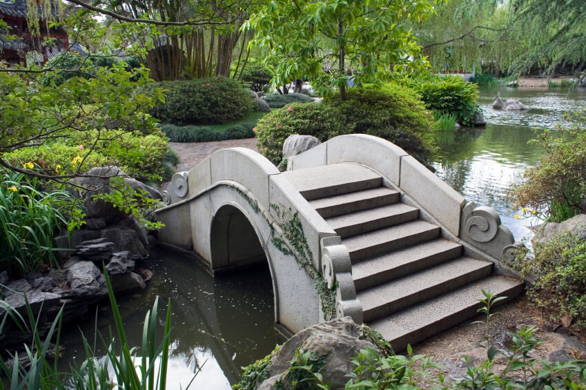An ornate concrete bridge across a wide but shallow and slow-moving creek. This bridge connects two sections of an expansive, lush garden. An ornate concrete bridge across a wide but shallow and slow-moving creek. This bridge connects two sections of an expansive, lush garden.