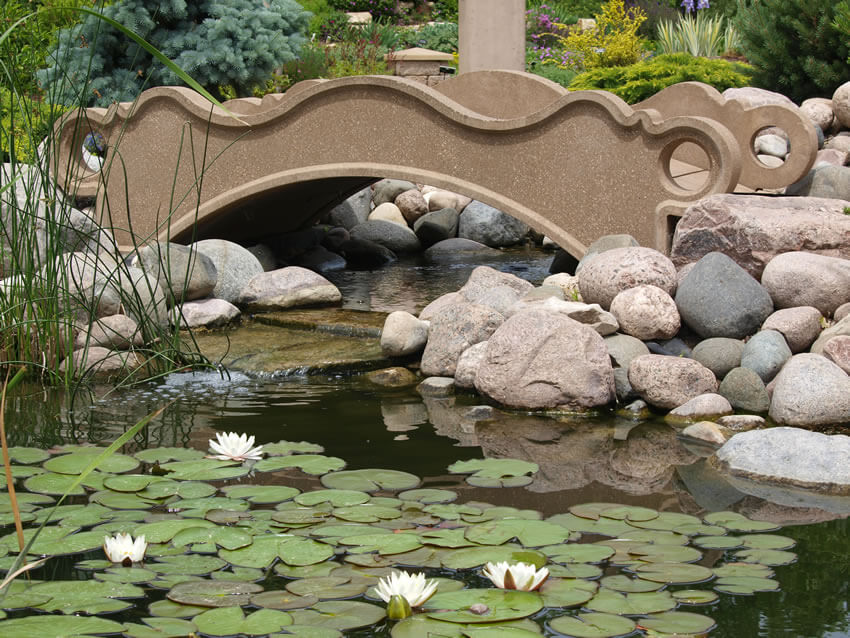 An ornate stone footbridge over a small creek leading into a pond. Stones piled up on either bank keep the bridge stead and in place. An ornate stone footbridge over a small creek leading into a pond. Stones piled up on either bank keep the bridge stead and in place.