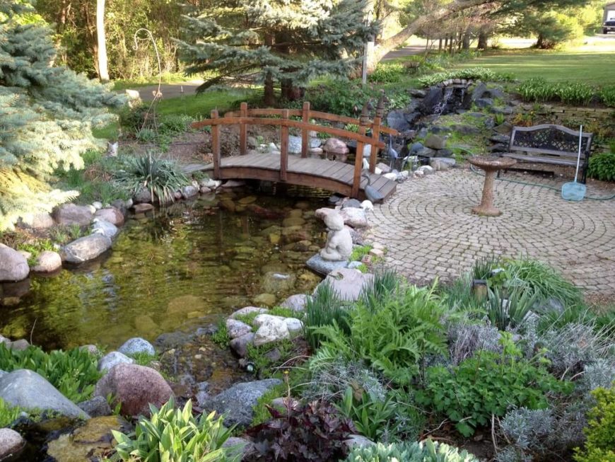 This simple Asian style footbridge leads from a circular patio to a small trail through the rest of the garden over a small pond fed by a stream. This simple Asian style footbridge leads from a circular patio to a small trail through the rest of the garden over a small pond fed by a stream.