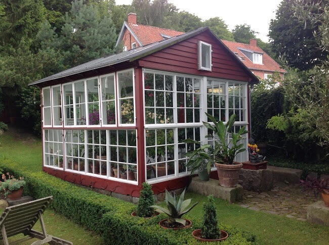 An adorable red and white greenhouse with a small stone walkway out front. The greenhouse is separated from the rest of the yard by a small, ankle-high hedge An adorable red and white greenhouse with a small stone walkway out front. The greenhouse is separated from the rest of the yard by a small, ankle-high hedge