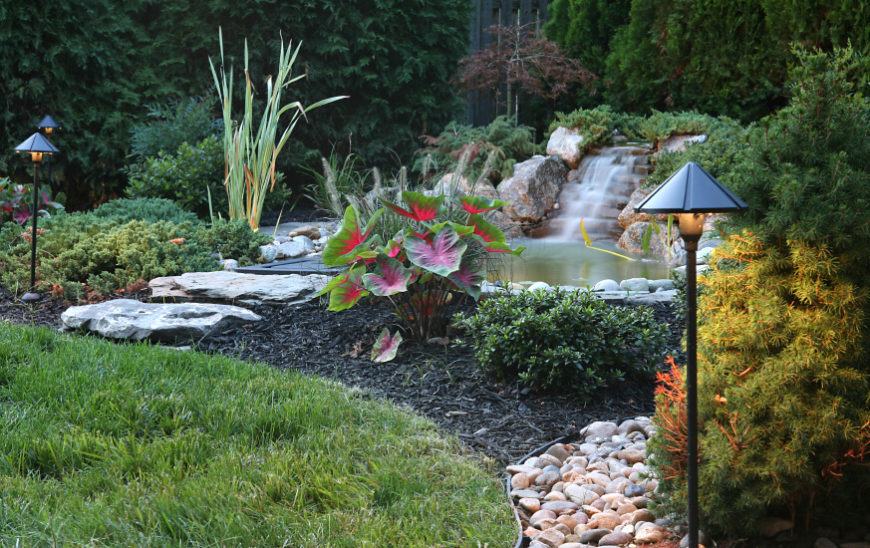 A large stone waterfall flows into a backyard pond with stepping stones leading up to the bank. A large stone waterfall flows into a backyard pond with stepping stones leading up to the bank.