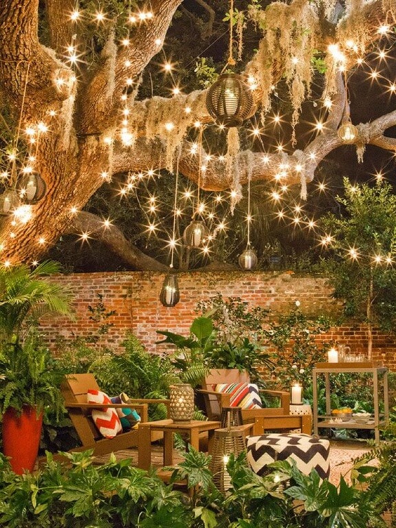 This space feels magical with the addition of lights and globes hanging from the gnarly branches of a willow tree. The adirondack chairs are nestled on a garden patio and accented with brightly patterned prints. This space feels magical with the addition of lights and globes hanging from the gnarly branches of a willow tree. The adirondack chairs are nestled on a garden patio and accented with brightly patterned prints.