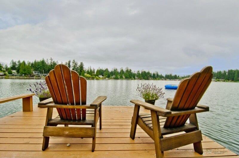 A set of adirondack chairs sit at the end of a dock with two planters filled with lavender. They look out over a pristine, calm lake. A set of adirondack chairs sit at the end of a dock with two planters filled with lavender. They look out over a pristine, calm lake.