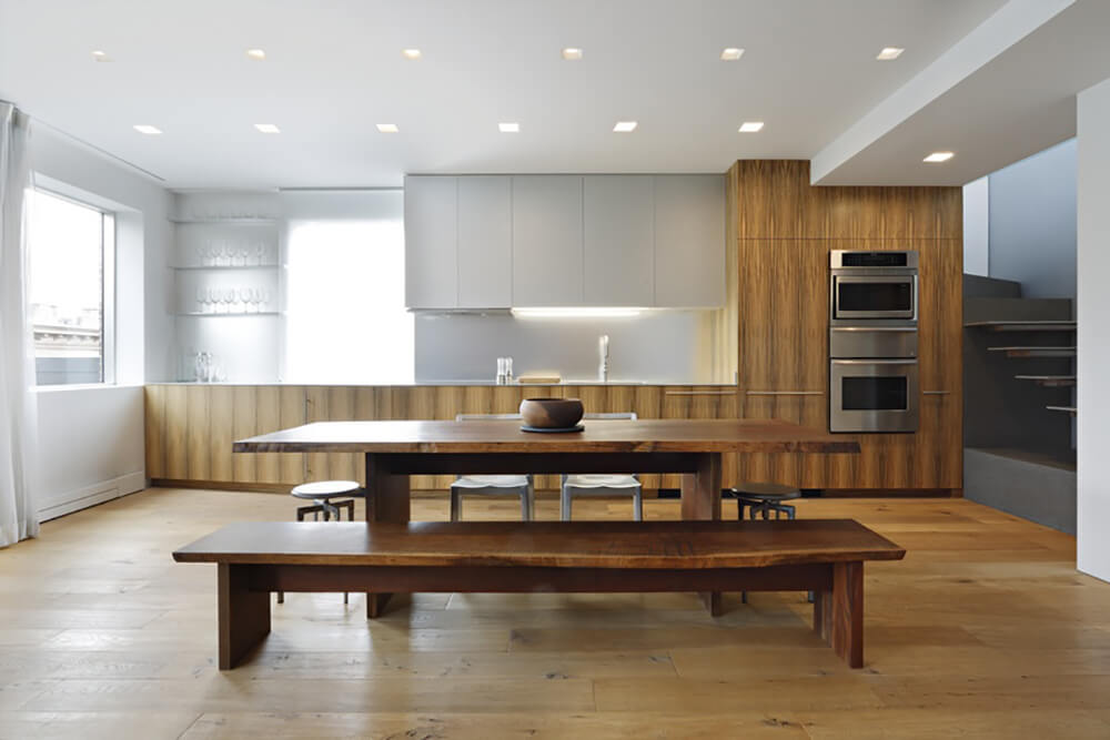 Simple kitchen with natural wood and white cabinets, floating shelves and stainless steel double ovens facing the wooden rectangular dining table and illuminated by square recessed lighting.