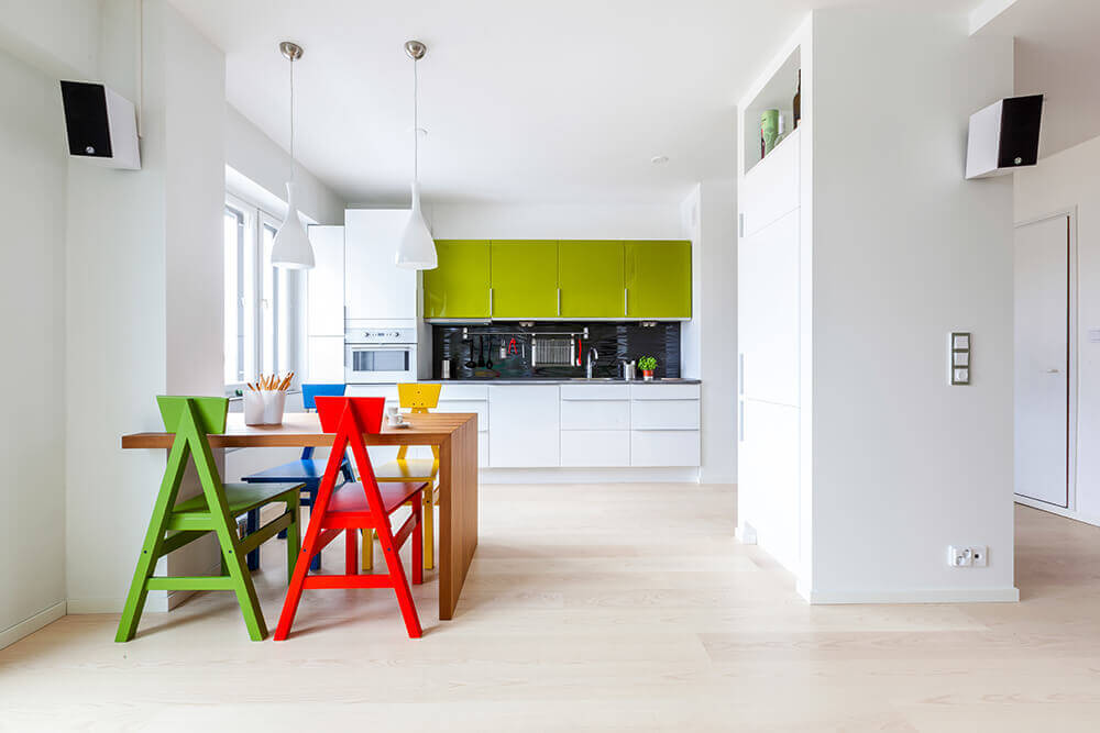 Beautiful white kitchen with black backsplash in the middle of glossy green and white cabinets over light hardwood flooring. A pair of white pendant lights hung over a small wooden kitchen table surrounded by unique and colorful wooden chairs.