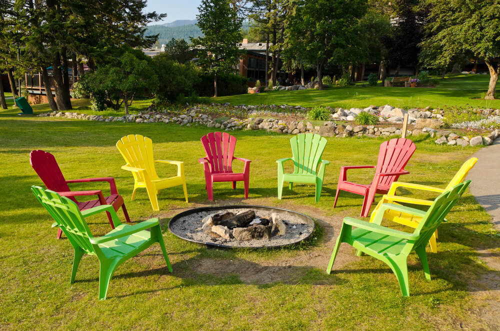 Dug in round fire pit with sand on grassy area surrounded by colourful Adirondack chairs. Dug in round fire pit with sand on grassy area surrounded by colourful Adirondack chairs.