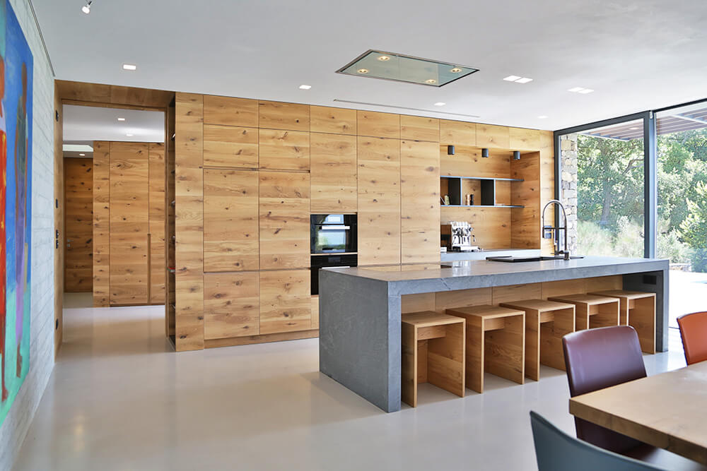 Dine-in kitchen with a gray island bar and natural wood cabinetry fitted with black double wall oven and floating shelves. It has smooth white flooring and floor to ceiling windows that bring plenty of natural light in.