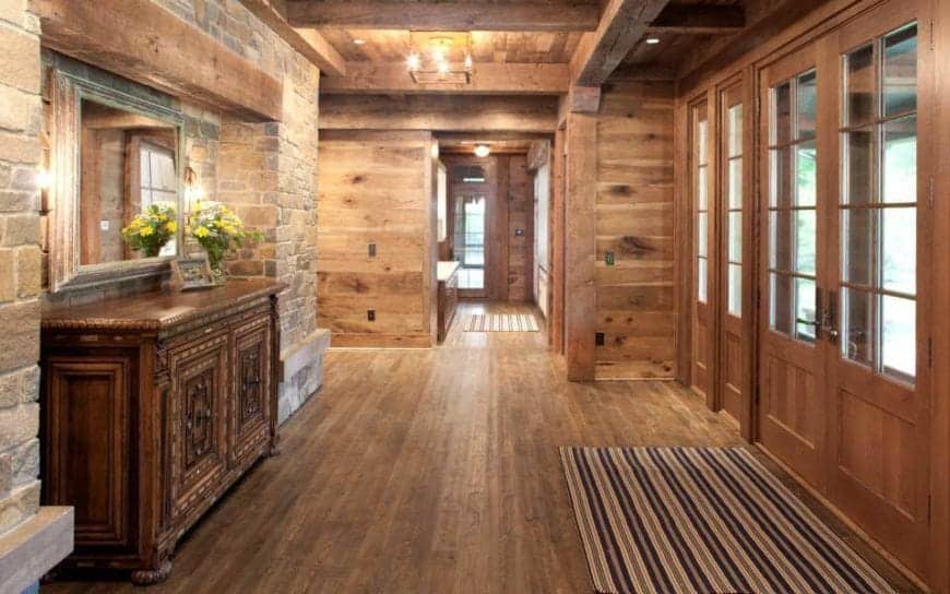 This charming foyer has set of double doors with a wooden finish and framing to its glass panels that brings in an abundance of natural lights to the hardwood flooring, wooden ceiling with exposed beams and the red brick wall across from the entrance.