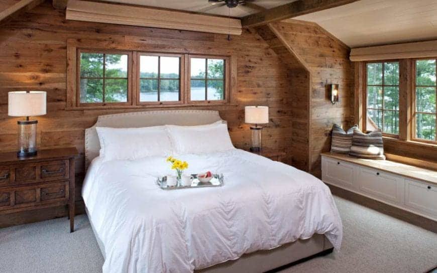 This is a charming Rustic-style bedroom with a light gray bed that has white sheets standing out against the gray carpeted flooring and the wooden walls. By the bed is a lovely sitting area with a built-in wooden bench by the window.