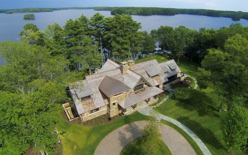 This aerial shot of the home allows for a better view of the surrounding lush greenery of the landscape that suits the lakeside terrain. The charming home is surrounded by well-maintained grass and tall trees. There is also on tall tree in the middle of the circular driveway in front of the house.