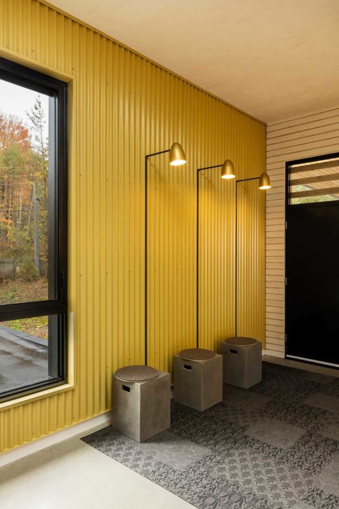This simple Industrial-style entry hall has a gray patterned area rug on its off white flooring. This is brightened by the delightful yellow hue of the walls made of metal and the wall-mounted lamps attached to it over three small built-in concrete stools.