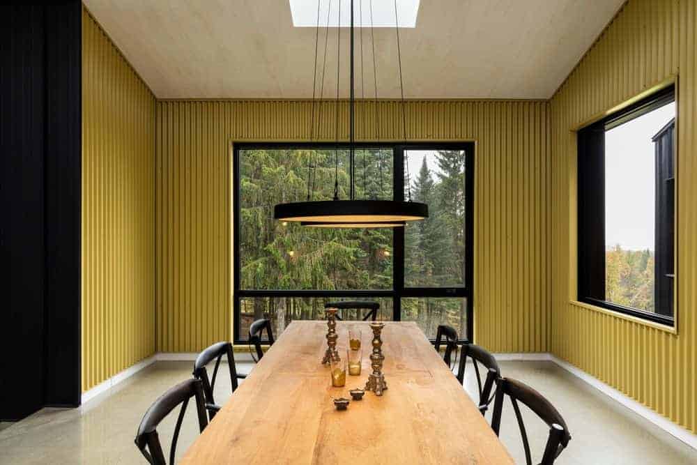The long rectangular wooden table stands out against the rest of the dining room that has cheerful yellow metal walls adorned with large glass windows that have contrasting black frames. These matches with the black wooden chairs that surround the table.