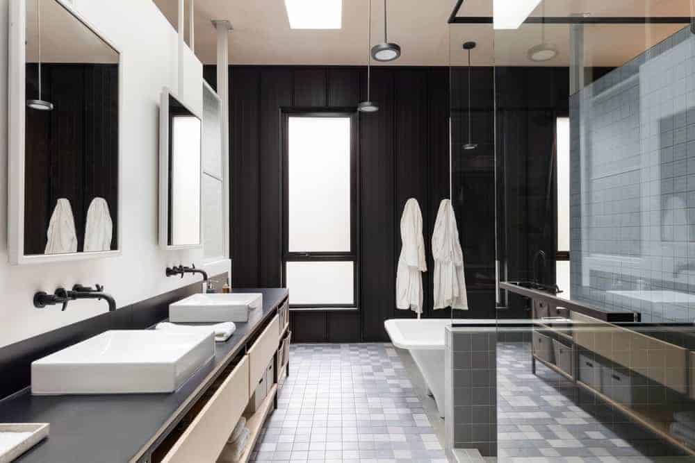 This modern industrial-style bathroom has gray flooring tiles that complement the black wall with a frosted glass window on the far end that matches with the countertop of the two-sink vanity with matching black fixtures.