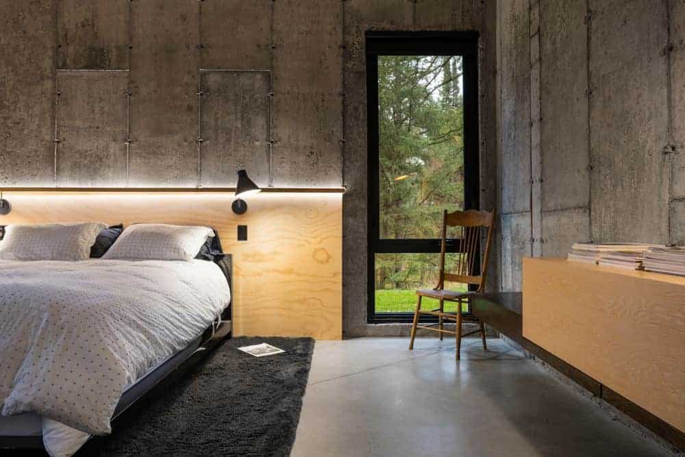 The concrete flooring that is covered with a black area rug matches with the upper layer of the gray concrete walls. The lower half of it is dominated by light wooden structures like the headboard and the floating cabinet on the adjacent wall.