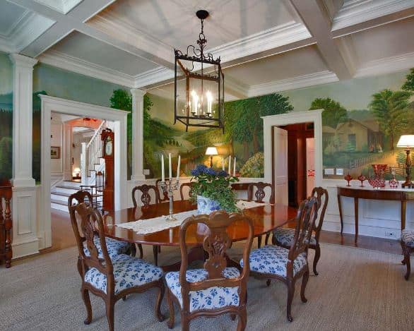 This elegant dining room has a white coffered ceiling that has a lantern-like pendant light over the oval dark wooden dining table and its wooden chairs contrasted by their white and blue seat cushions. This setup is given a nice background of walls dominated by a riverside farm mural.