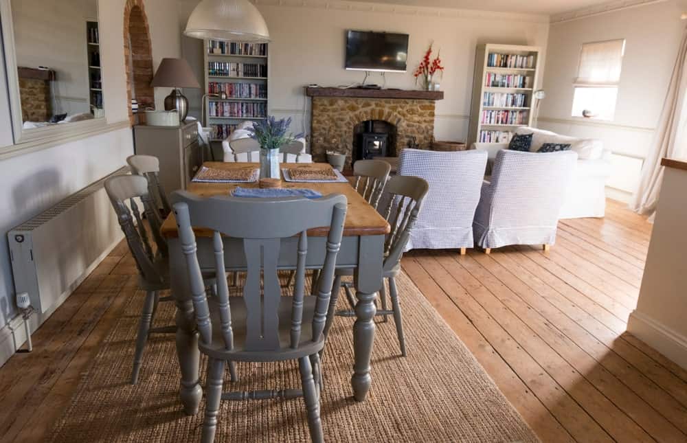 This dining set has beautiful light gray wooden chairs with a classic design matching the legs of the rectangular wooden dining table. This is placed on a woven area rug over the hardwood flooring by the living room that has a fireplace.