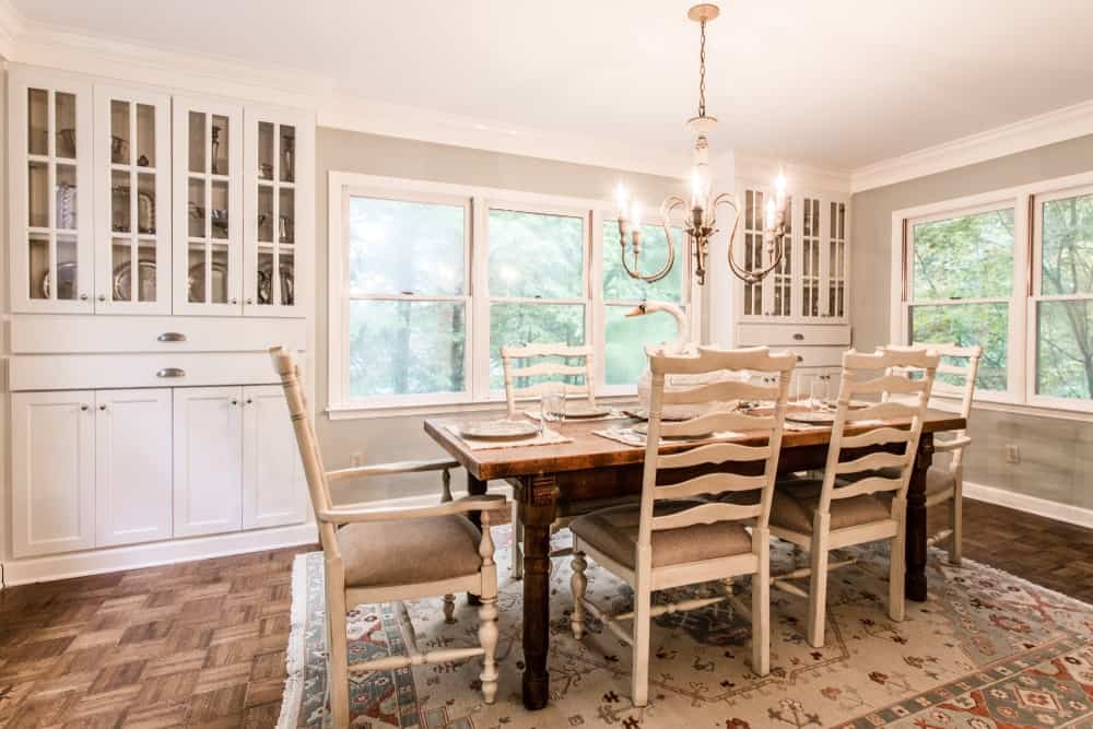 The lush green trees outside the glass windows adds a bit of color to the background of the wooden dining table that has contrasting white wooden chairs. These match with the light hue of the patterned area rug that is illuminated by the charming chandelier.