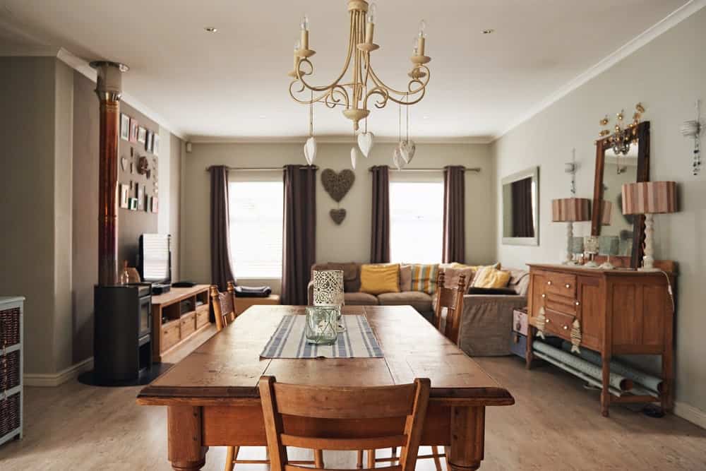 The lovely chandelier hanging from the white ceiling has a beige tone to its intricate structure. This adds a bit of flair to the simple dining table and its dining chairs that match the hardwood flooring and the cabinets that stand out against the light gray walls.