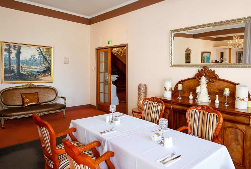 The armchairs with redwood frames and striped colorful cushions stand out against the white table cloth of the rectangular dining table that contrasts the dark gray area rug over the terracotta flooring complemented by the beige walls.