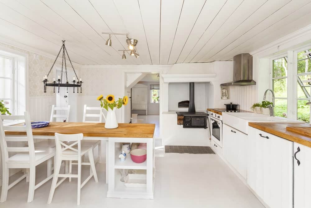 This is a bright and predominantly white dining area inside the kitchen with the same white shiplap ceiling and white flooring. The brightness is subdued by the wooden tabletop of the dining table and the black farmhouse-style chandelier hanging above it.