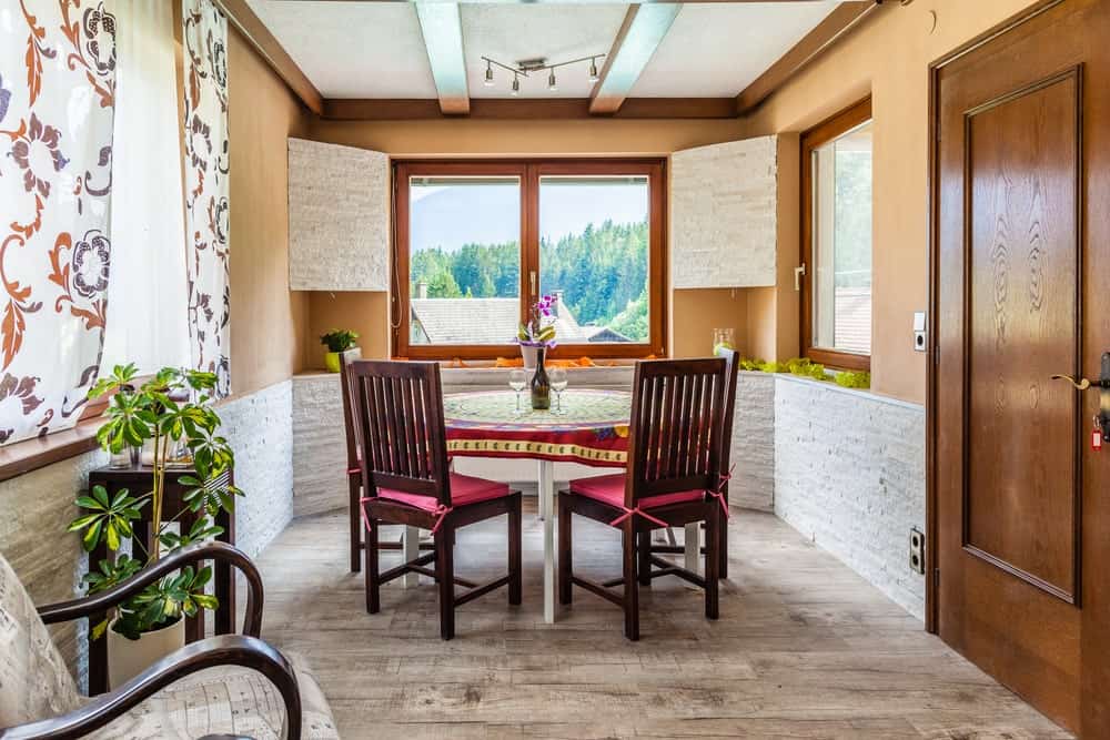 This small dining room is surrounded by orange walls with windows to give a sense of space. The walls have a textured white stone wainscoting that provides a nice contrast to the dark wooden slat back dining chairs of the white round table with a colorful table cloth.