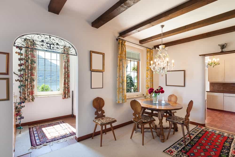 The majestic white chandelier elevates this small dining area at a corner by the window. It has a simple round wooden dining table that can seat four people on its wooden chairs that has floral seat cushions matching the area rug.