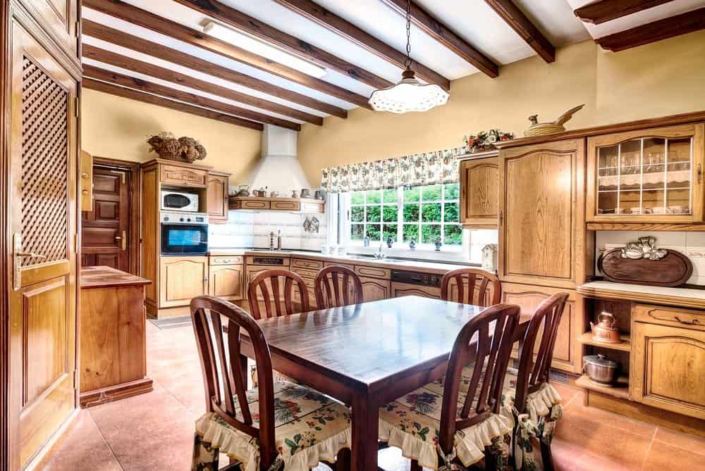 This is an informal dining area inside the kitchen that has terracotta flooring tiles. This complements the dark wooden table and its chairs of the same hue that is accented with floral seat cushions that matches the pendant light hanging from the exposed wooden beams of the ceiling.