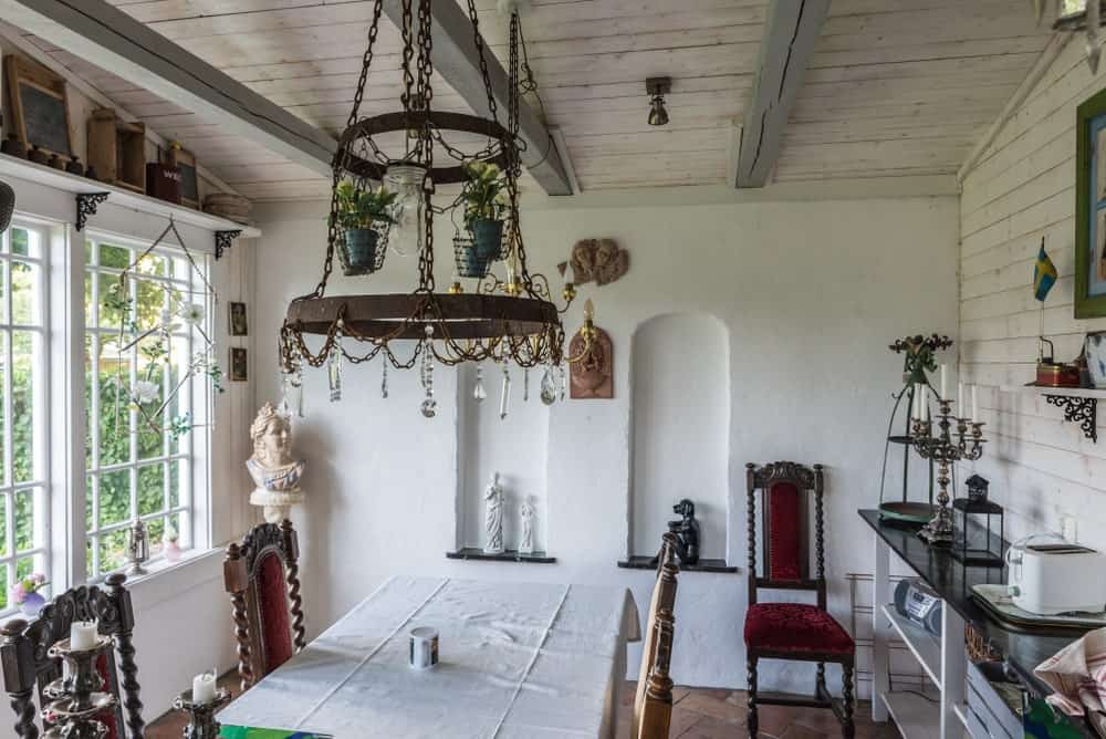 This Country-style dining room has a rustic charm to its wooden walls and wooden ceiling with exposed beams supporting a decorative pendant light that has crystals and potted plants hanging over the rectangular dining table and its classic wooden chairs.