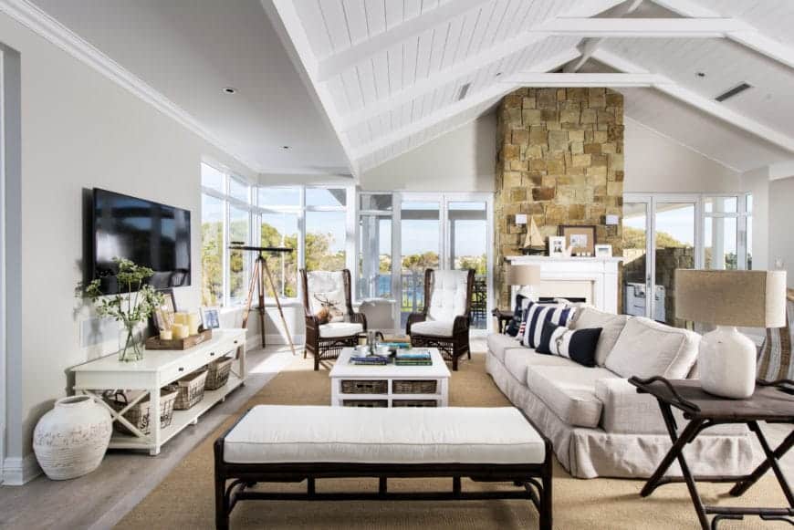 This is a large inviting living room that treats its guests with a bright comfort. It has a high white cathedral ceiling with exposed beams matching the window frames that bring in natural lights to the large beige sofa over a woven area rug.